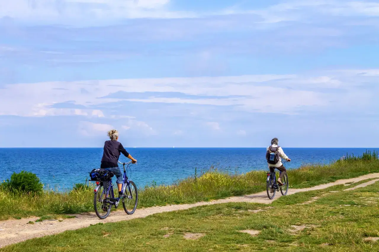Dwie osoby jadą rowerami po malowniczej trasie rowerowej wzdłuż Bałtyku. Błękitne niebo i morze tworzą idealne tło dla tej spokojnej przejażdżki.
