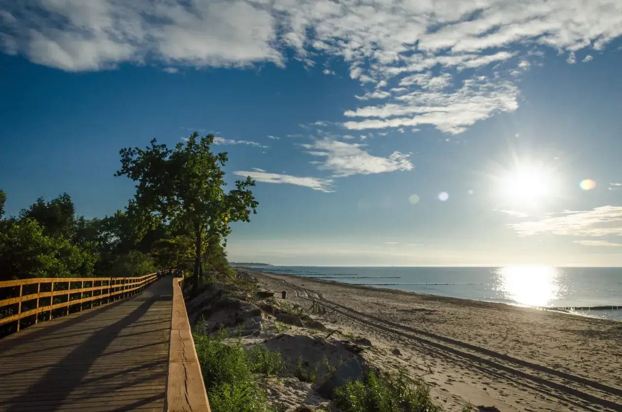 Drewniana ścieżka prowadzi wzdłuż piaszczystej plaży. Słońce odbija się w spokojnym morzu. Idealna trasa rowerowa wzdłuż Bałtyku.