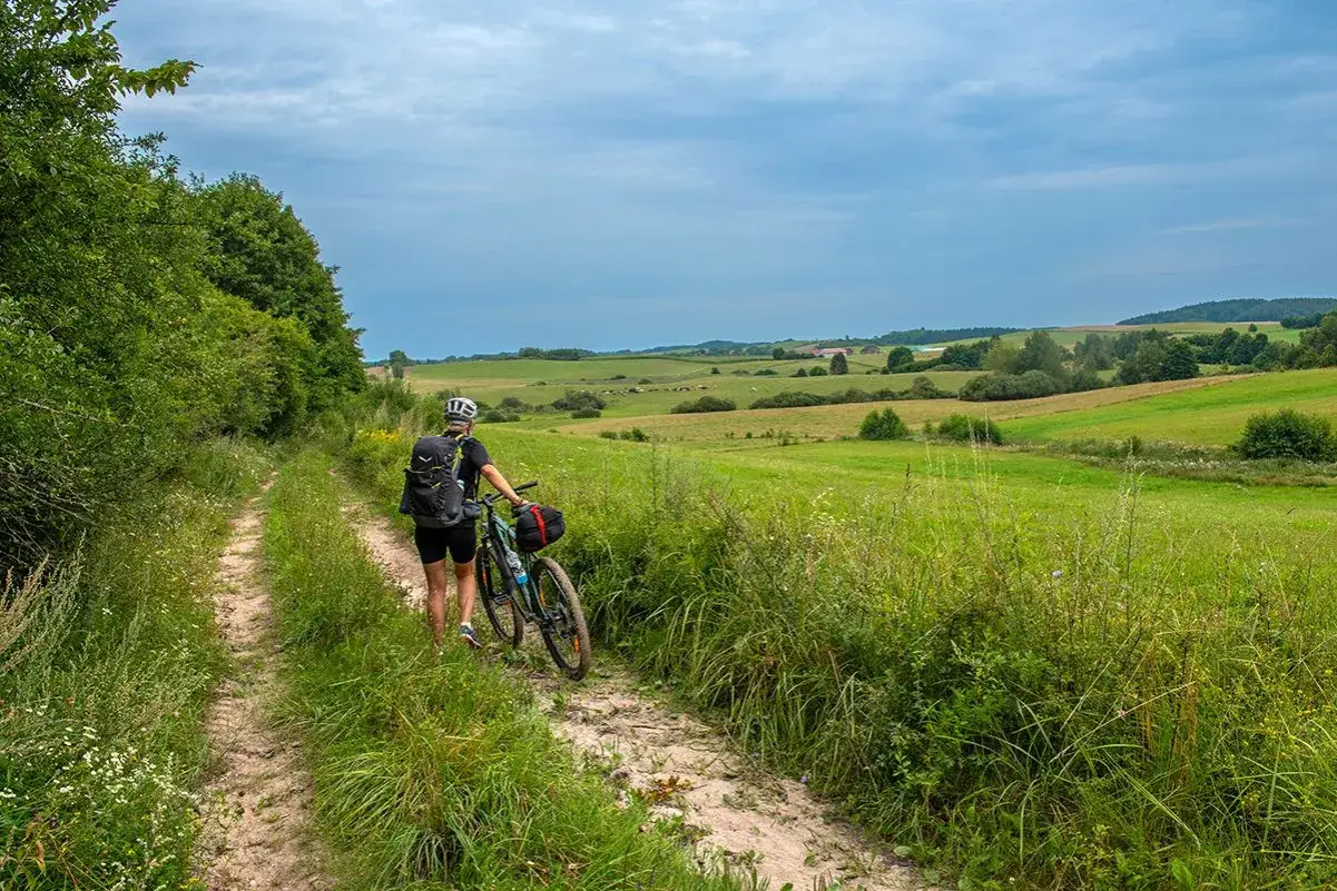Kobieta z rowerem na szutrowej drodze, odkrywa malownicze trasy rowerowe Mazury. Zielone pola i pagórki tworzą sielski krajobraz.