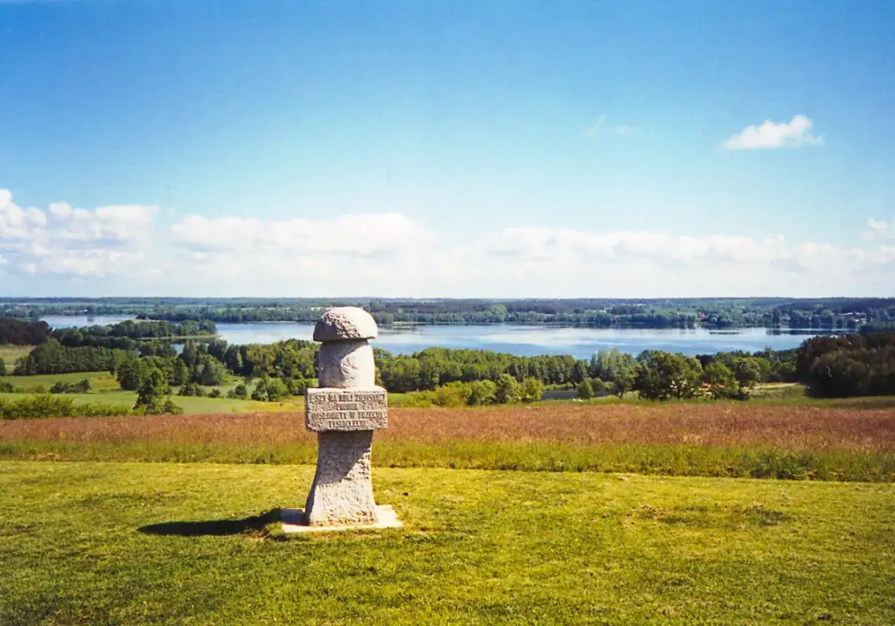Kamienny obelisk na wzgórzu, z widokiem na jeziora i lasy. To początek szlaku stu jezior, zapraszającego do odkrywania piękna Mazur.