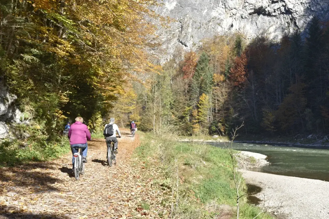 Jesień na trasach rowerowych w Łódzkiem. Ludzie na rowerach jadą ścieżką wśród kolorowych liści i drzew, obok rzeki i skalnej ściany.