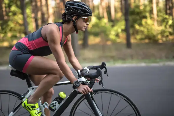 Kobieta w kasku i okularach jedzie na rowerze szosowym przez las. Widać jej nogi w sportowym stroju i zegarek na ręce. To dowód, że jazda na rowerze efekty po miesiącu są widoczne!