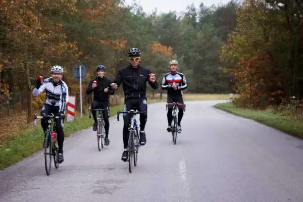 Peleton kolarzy jedzie drogą wśród jesiennych drzew. Czy to wyścig, czy wspólna przejażdżka?